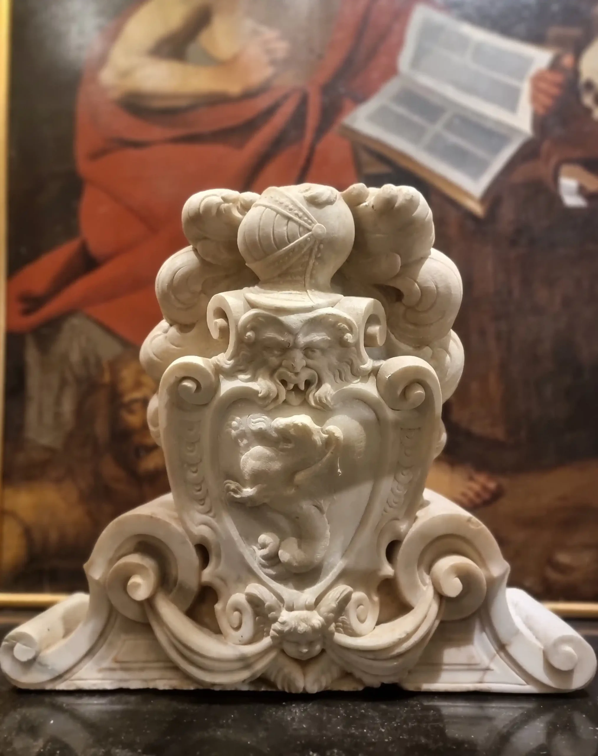 Scultura in marmo bianco raffigurante un elmo, un volto grottesco e uno scudo con un leone rampante, su sfondo sfocato di un dipinto antico con figure drappeggiate in rosso.